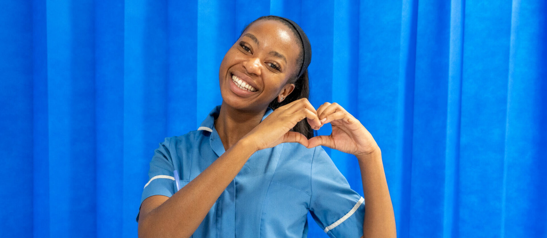 Nurse in-front of a blue curtain doing a heart sign with her hands